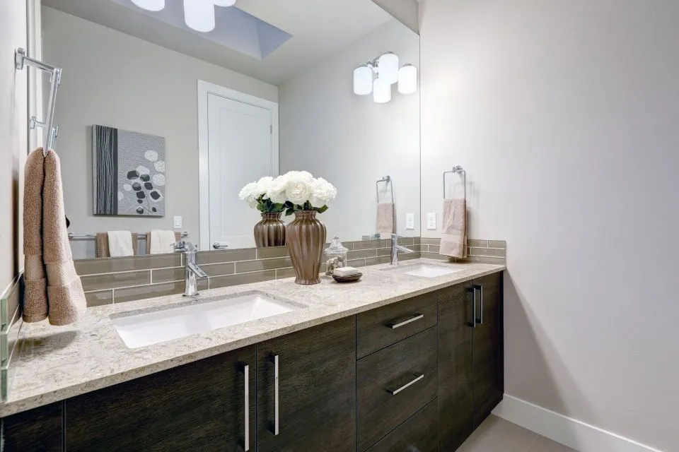 Clean bathroom vanity with dark wood double-sink cabinet, light granite countertop, large mirror, and white floral arrangement